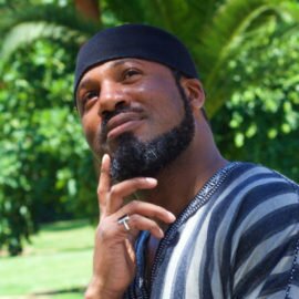 Ahmad Ikhlas in a black kufi, thoughtful, against green palms